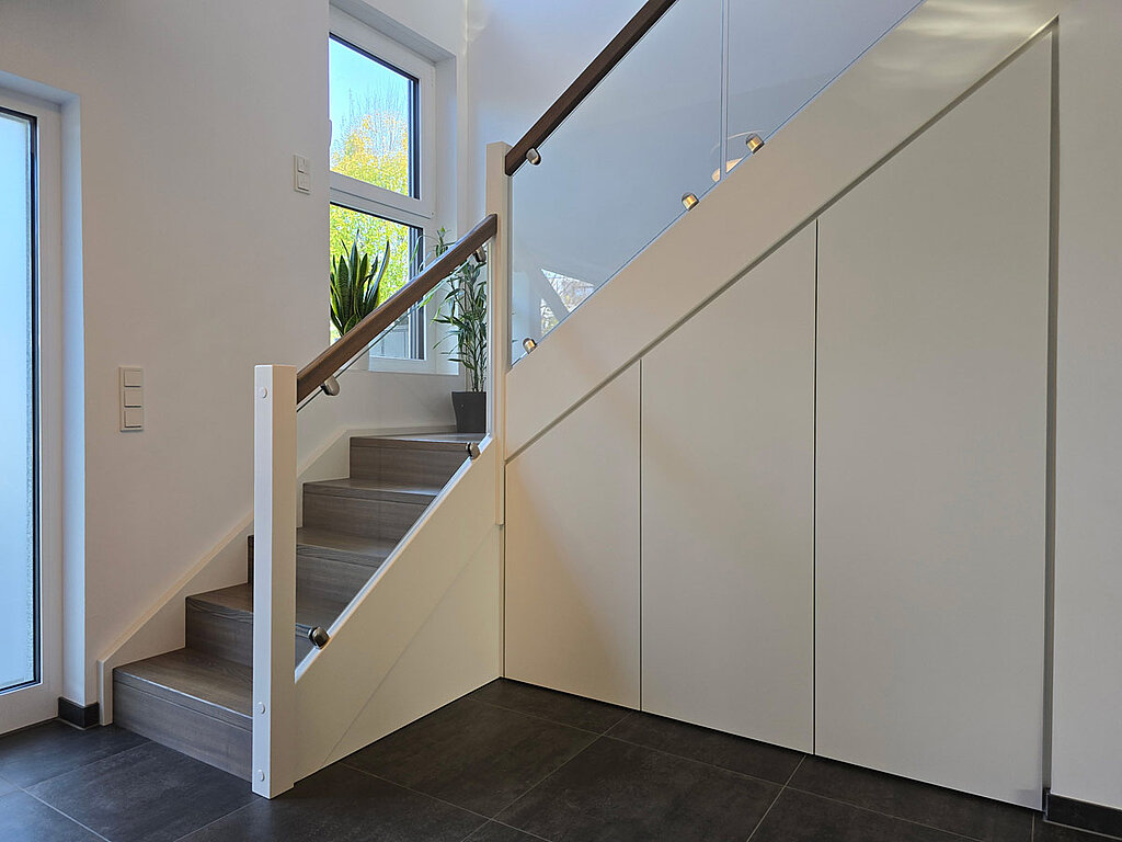 Der oft ungenutzte Raum unter der Treppe wurde hier in ein vollwertiges Stauraumwunder verwandelt. Die maßgefertigten Schranktüren folgen präzise der Unterseite der Treppe. Durch das grifflose Design in Weiß tritt der Schrank optisch zurück und überlässt der Treppe mit ihren dunklen Stufen und dem modernen Glasgeländer die Bühne.