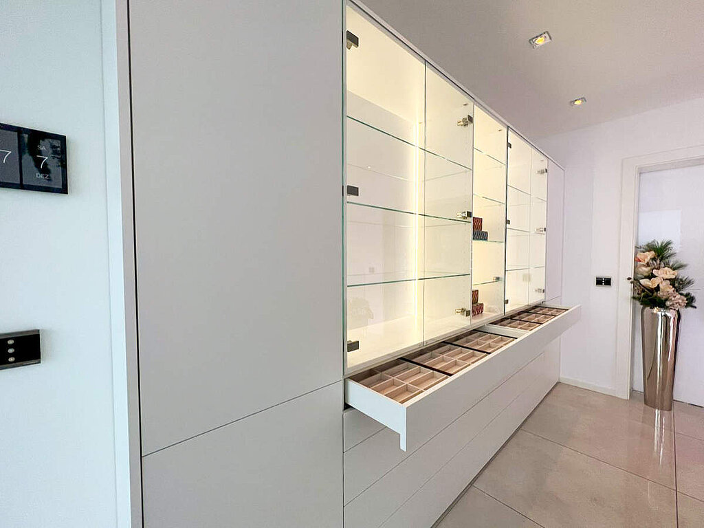Dieses elegante Einbaumöbel kombiniert eine beleuchtete Glasvitrine mit darunterliegenden Schubladenelementen. Ein besonderer Fokus liegt auf der Funktionalität: Die flachen Auszüge sind mit einer individuellen Facheinteilung ausgestattet, die sich ideal für die geordnete Aufbewahrung von Brillen oder Accessoires eignet.
