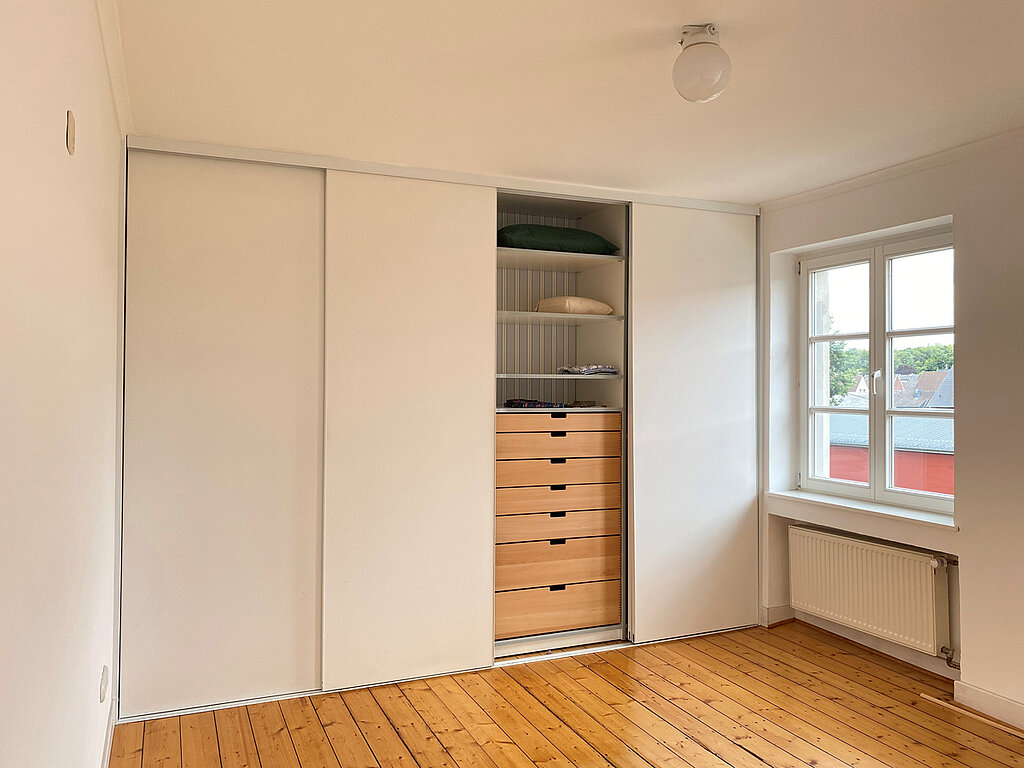 Auf dem ersten Blick ganz unscheinbar - Durch die weißen Schiebetüren wirkt der Einbauschrank wie eine vierte Wand im Raum. Hinter den Türen verbirgt sich aber reichlich Stauraum, der, gerade in kleineren Wohnungen, dringend benötigt wird.