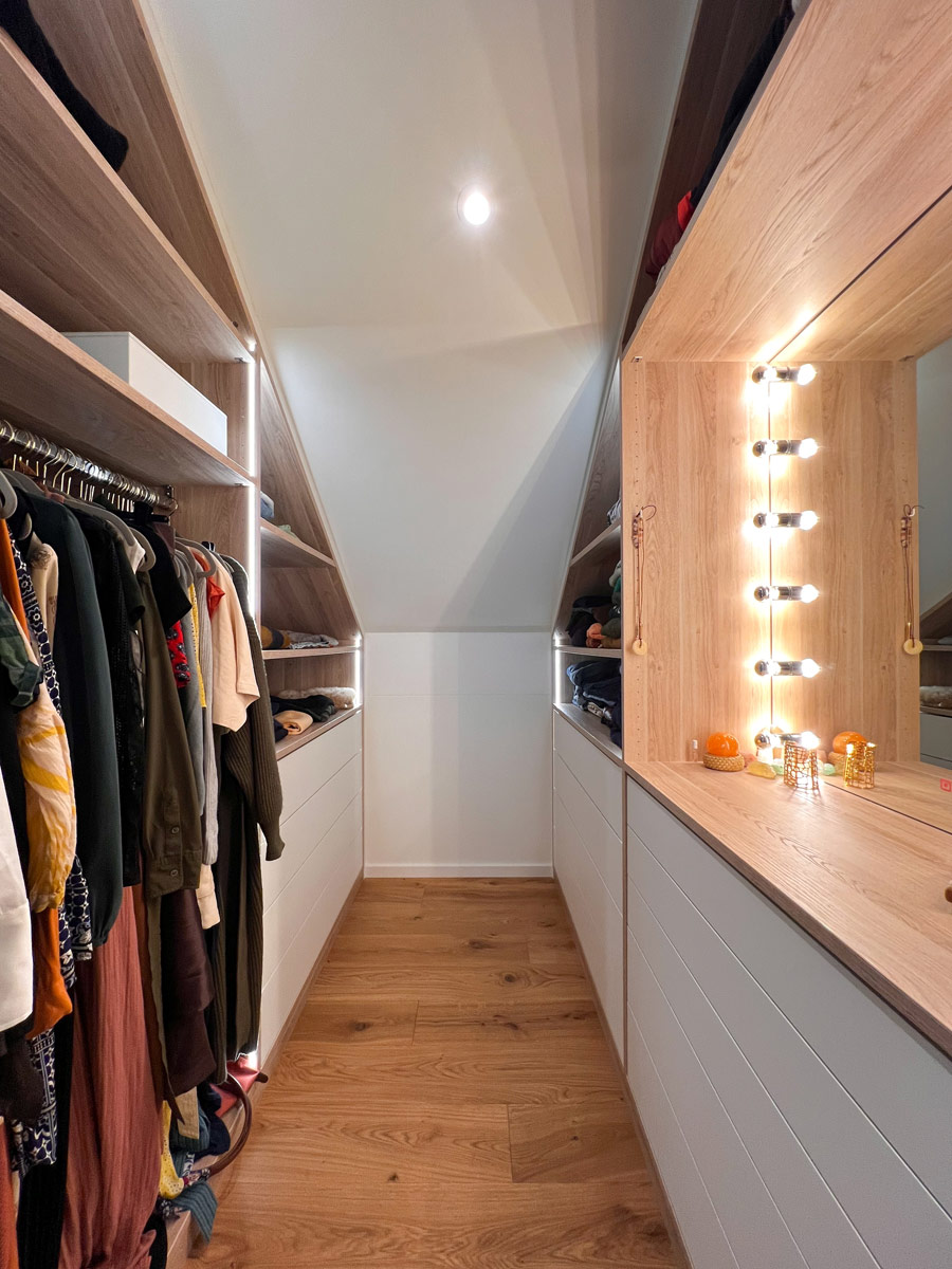 Dieses Projekt zeigt, wie Dachschrägen effizient genutzt werden können. Die Einbauschränke folgen exakt dem Neigungswinkel der Decke. Auf der rechten Seite wurde ein beleuchteter Schminkplatz mit großem Spiegel direkt in die Schrankzeile integriert. Die Kombination aus weißen Fronten und Oberböden in Holzoptik sorgt für eine helle, einladende Raumwirkung trotz der begrenzten Deckenhöhe.