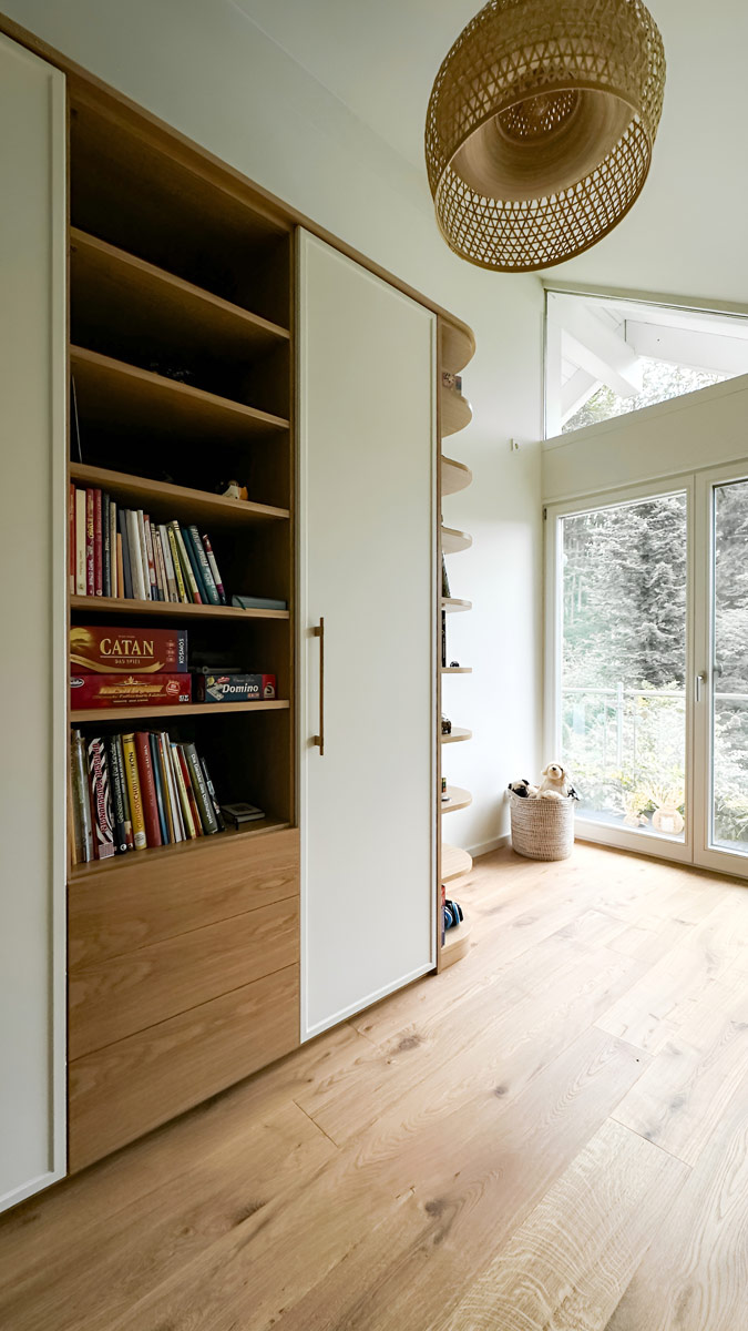 Dieses deckenhohe Einbaumöbel verbindet offenes Design mit funktionalem, geschlossenem Stauraum. Die Kombination aus weißen Fronten und Korpus-Elementen in heller Eiche schafft eine freundliche, skandinavisch angehauchte Atmosphäre. Das Herzstück bildet das offene Bücherregal auf der linken Seite. Die rechte Seite ist mit klassischen Drehtüren ausgestattet, die sich über markante Holzgriffe öffnen lassen und den geschlossenen Stauraum verbergen. Ein besonderes handwerkliches Detail sind die abgerundeten Abschlussregale zur Fensterseite hin, die einen sanften Übergang in den Raum schaffen.